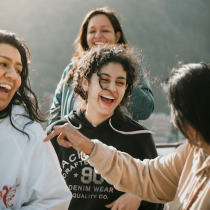 Four women wearing Cultivating Joy apparel are outdoors, laughing and smiling together under the sun, creating a warm and joyful atmosphere.